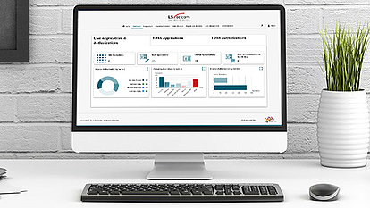 A desktop computer on a white desk displaying a spectrum management dashboard with charts and graphs. The screen shows user applications, TDMA applications, and authorizations with pie charts, bar graphs, and summary statistics. A plant in a white vase and a wireless mouse are placed next to the keyboard.